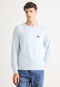 Light blue sweater made of soft knit fabric, featuring a round neckline, long sleeves, and a small black circular logo on the chest.
