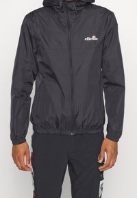 Coupe-vent léger noir avec fermeture éclair à l'avant, poignets et ourlet élastiques. Doté d'un petit logo blanc sur la poitrine et d'une capuche.