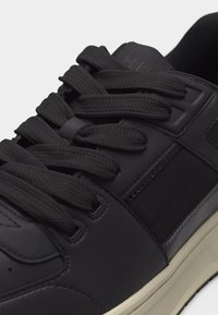 Gros plan sur une basket noire avec des lacets noirs épais, des panneaux latéraux texturés et une semelle blanche sur un fond clair.