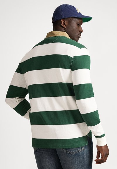 Muž oblečený v zeleno-bielom širokom pruhovanom polokošeli s dlhým rukávom, modrých rifliach a tmavomodrej šiltovke, otočený mierne od kamery.