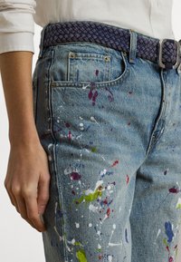 Zila džinsu bikses ar rozā, zaļu un baltu krāsas pilieniem uz priekšpuses. Redzama ir pītā purpura josta ar sudraba sprādzi.