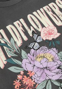 T-shirt nera con il testo "Wildflowers" in rosa chiaro, accompagnato da illustrazioni floreali e una farfalla in blu e nero, su un tessuto texturizzato.