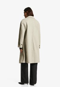 Osoba s dlhými tmavými vlasmi má na sebe svetlo béžový kabát do kolien, čierne široké nohavice a čierne lodičky, stojí chrbtom na bielom pozadí.