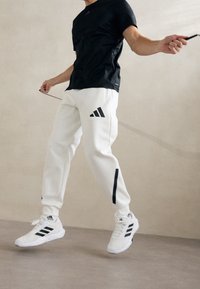 Bílé sportovní kalhoty s černými akcenty a logem tří pruhů, spojené s černým tričkem s krátkým rukávem a bílými teniskami, uvnitř.