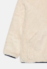 Giacca in pile di colore crema con una texture morbida e pelosa. Presenta un bordo blu navy sui polsini e una tasca a marsupio sul davanti.