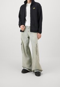 Person med svart Nike zip-up jacka, vit t-shirt, ljusgrå vida cargobyxor och svarta platåskor som står mot en enfärgad bakgrund.