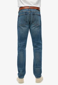 Jeans de mezclilla azul con corte recto, que cuentan con un cinturón marrón, dos bolsillos traseros y un desgaste sutil. Zapatos con suela de goma visibles en la parte inferior.