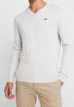 Pull ras de cou gris clair en maille douce, avec des poignets et un ourlet côtelés, et un petit logo rouge sur la poitrine.