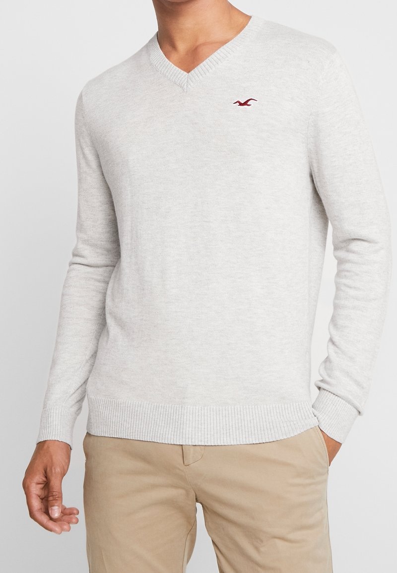 Pull ras de cou gris clair en maille douce, avec des poignets et un ourlet côtelés, et un petit logo rouge sur la poitrine.