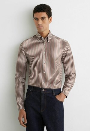 Młody mężczyzna w brązowo-białej pionowo prążkowanej koszuli zapinanej na guziki, wsadzonej w ciemnoniebieskie dżinsy, stoi na tle jednolitej białej ściany.