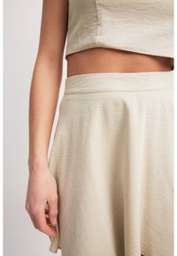 Beige kahden osan asu, joka koostuu lyhyestä crop-topista ja A-linjaisesta hameesta. Kangas on pehmeä ja tekstuuri on hienovarainen kiilto.