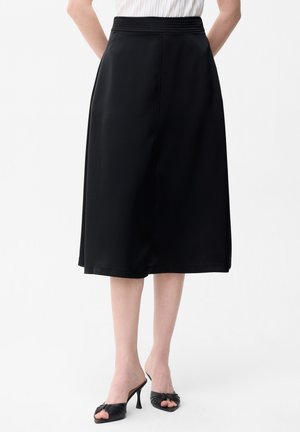 Schwarzer Midirock mit hohem elastischem Bund, getragen mit schwarzen offenen, hochhackigen Mules und einem weißen, in den Rock gesteckten ärmellosen Top.