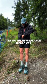 Black waterproof jacket with red accents, paired with black shorts. Wearing teal New Balance Hierro V9S shoes with white socks on gravel trail.