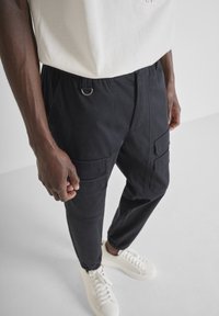 Man in zwarte cargobroek met zijzakken en witte sneakers, staande tegen een effen lichte achtergrond.