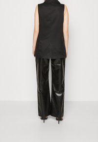 Femme portant un blazer noir sans manches, un pantalon large noir brillant et des sandales à talons hauts noires, debout devant un fond uni.