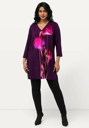 Paarse tuniek met een diepe V-hals en 3/4 mouwen, voorzien van een levendige bloemenprint in roze en rood. Gecombineerd met zwarte slim-fit broek.