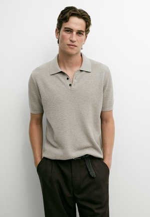 Giovane uomo con capelli scuri indossa una polo beige a maniche corte con texture e pantaloni neri con cintura intrecciata, in piedi contro un muro bianco.
