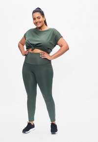 Olijfgroene cropped top met een knoopdetail in de taille, gecombineerd met olijfgroene high-waisted leggings. Model draagt zwarte sportschoenen.