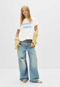 T-shirt en coton blanc avec du texte bleu, associé à un pantalon en jean large délavé bleu clair. Un pull jaune drapé sur les épaules.
