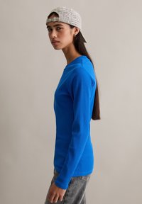 Niebieska prążkowana bluzka z długim rękawem i okrągłym dekoltem, zestawiona z szarymi dżinsami. Dopełnieniem stroju jest czapka w kratę, dodająca casualowego akcentu.