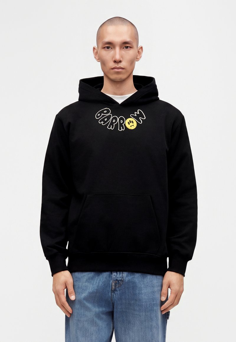 Homme portant un sweat à capuche noir avec le texte "PARROW" et un visage triste jaune, associé à un jean bleu, debout devant un fond clair uni.
