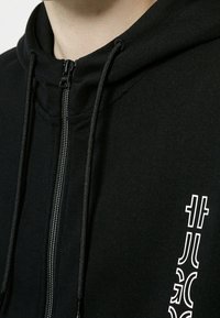 Felpa nera con zip realizzata in misto cotone. Presenta un colletto alto, lacci e un logo bianco sul lato sinistro. Texture liscia.