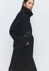 Abrigo corto de lana azul marino con cuello alto, bolso de mano de cuero negro con largas flecos, llevado sobre una falda de tela oscura.