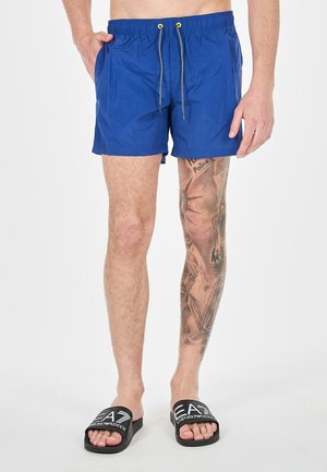 Costume da bagno blu con vita elastica, cordoncino regolabile e due tasche laterali. Indossato con infradito nere con logo.