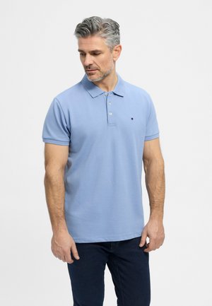 Un homme aux cheveux gris présente un polo bleu clair et un pantalon foncé sur un fond blanc uni.