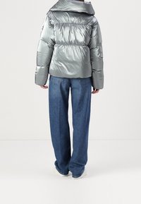Silver puffer jacket with a high collar, quilted texture, and elasticized waist, paired with loose-fit blue denim jeans and white sneakers.