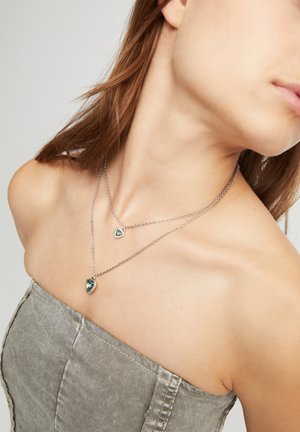 Collar de tonos plateados en capas que presenta dos colgantes en forma de corazón con acentos verdes. La cadena es fina y tiene una textura delicada.