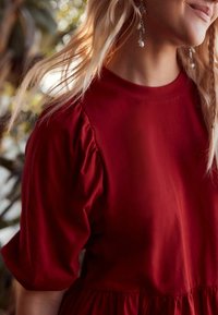 Robe rouge à manches courtes en tissu lisse. Caractéristiques : manches froncées et bas évasé avec un col rond, sur un fond flou.