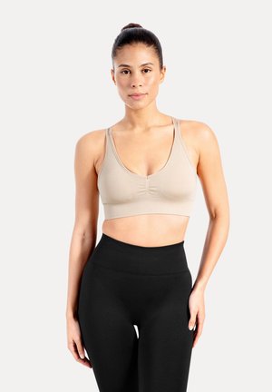 Mujer con el pelo recogido en un moño, vistiendo un sujetador deportivo beige y leggings negros de cintura alta, de pie contra un fondo claro y liso.