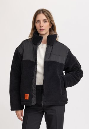 Vrouw die een zwarte fleecejas draagt met gevoerde schouders en kraag, witte trui en zwarte broek, staand met één hand in de zak.