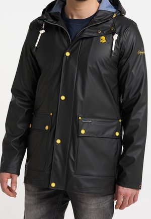 Hombre con chaqueta impermeable negra con botones amarillos, bolsillos frontales y capucha con cordones blancos sobre una camisa azul marino.