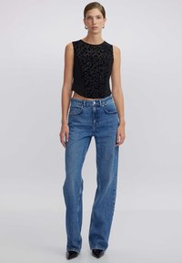 Top negro sin mangas con patrón de leopardo texturizado, combinado con jeans de pierna ancha de tiro alto en azul. La modelo posa frente a un fondo sencillo.