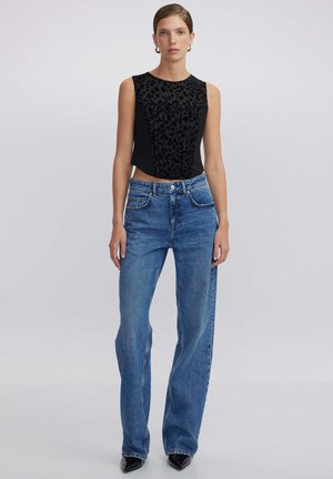 Schwarzes ärmelloses Top mit strukturiertem Leopardenmuster, kombiniert mit hoch taillierten blauen Weitbeinjeans. Das Model steht vor einem schlichten Hintergrund.