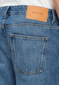 Bakfraust av blå denim jeans med baklomme og tanfarget lær merke med teksten "ONLY & SONS" sydd på midjebåndet.