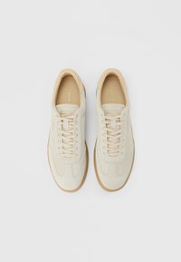 Poros šviesiai rudos zomšos apsiūti sportiniai batai su guminiu padais, žvelgiant iš aukščio ant baltos fono.