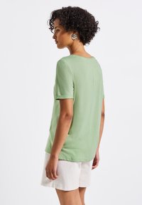 T-shirt vert à manches courtes avec un col rond, en tissu doux, coupe légèrement oversize, et un détail de couture unique au dos. Associé à un short beige.