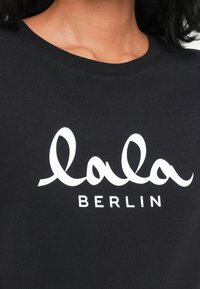 Melna kokvilnas T-krekls ar apaļu kakla izgriezumu, kuram ir liels balts grafiskais prints ar uzrakstu "lala BERLIN." Gluda tekstūra, īsās piedurknes.
