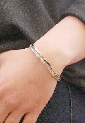 Gouden metalen armband met een gladde, gepolijste oppervlakte en een textuurband met heldere accenten, gedragen om een pols.