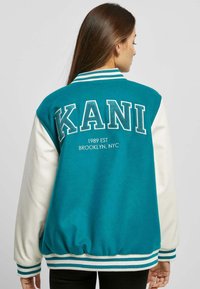 Veste de varsity teal avec des manchons et un ourlet blancs, poignets côtelés. L'arrière présente un texte brodé "KANI" et "1989 EST BROOKLYN, NYC."