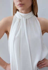 Haut blanc sans manches à col montant en tissu lisse, avec des plis doux au col et un design fluide. Des boucles d'oreilles créoles dorées sont visibles.