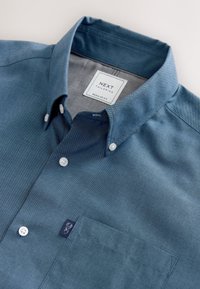 Chemise à manches courtes boutonnée en tissu texturé bleu marine, dotée d'un col pointu, de boutons blancs et d'une poche poitrine avec un logo.