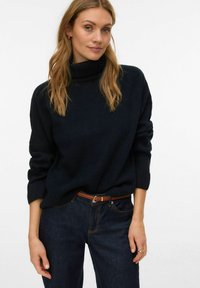 Tumši zils augstas apkakles džemperis ar vaļēju piegriezumu, ribotām aprocēm un smalku tekstūru, kombinēts ar tumšām augsta vidukļa džínsām un brūnu jostu.