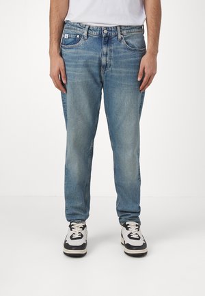 Homme portant un jean bleu délavé, des baskets blanches et noires, et une chemise blanche, debout devant un fond blanc uni.