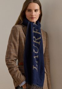 Marinblå halsduk av mjukt tyg, med kontrasterande ljusa bokstäver och fransar i ändarna, draperad över en rutmönstrad blazer.