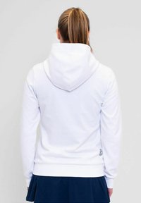 Sudadera blanca con un bolsillo tipo canguro y capucha con cordón, con dobladillo y puños acanalados. Combinada con una falda plisada azul marino.