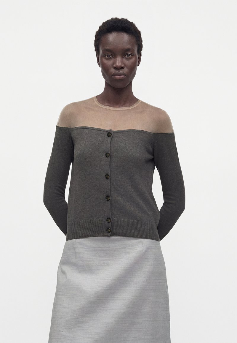 Woman with short curly hair wearing a buttoned charcoal cardigan with a sheer beige yoke and a light gray skirt, standing against a white background.
